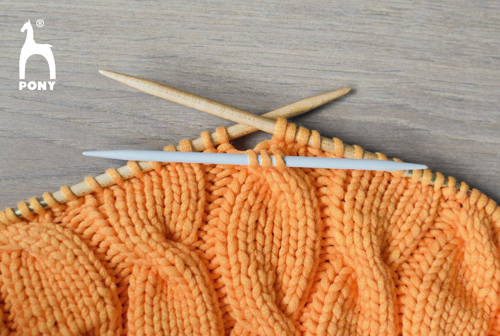 Knitted fabric with orange yarn and knitting needles on a wooden surface, featuring the Pony brand logo.