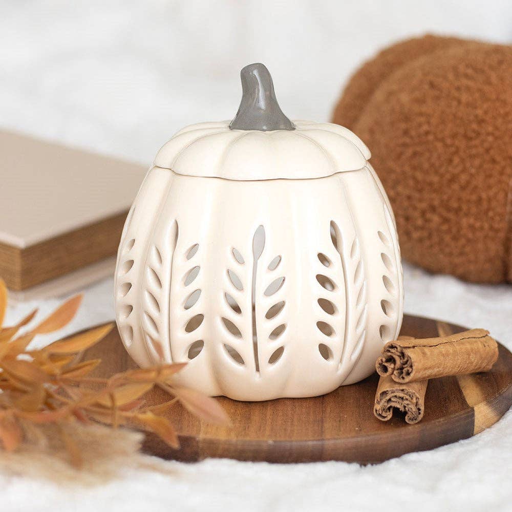 Ceramic pumpkin-shaped lantern with cut-out designs on a wooden surface with cinnamon sticks and leaves.