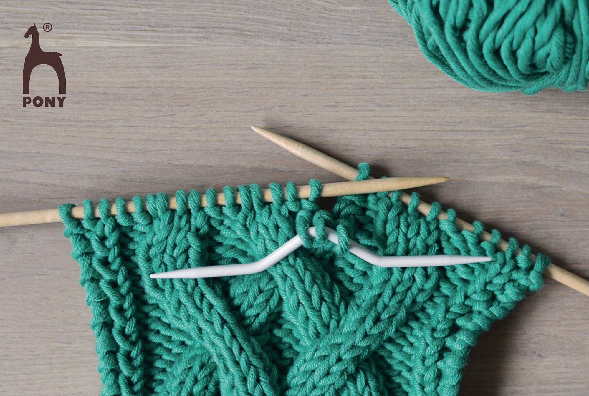 Green yarn being knitted with wooden needles on a wooden surface, featuring the Pony brand logo.
