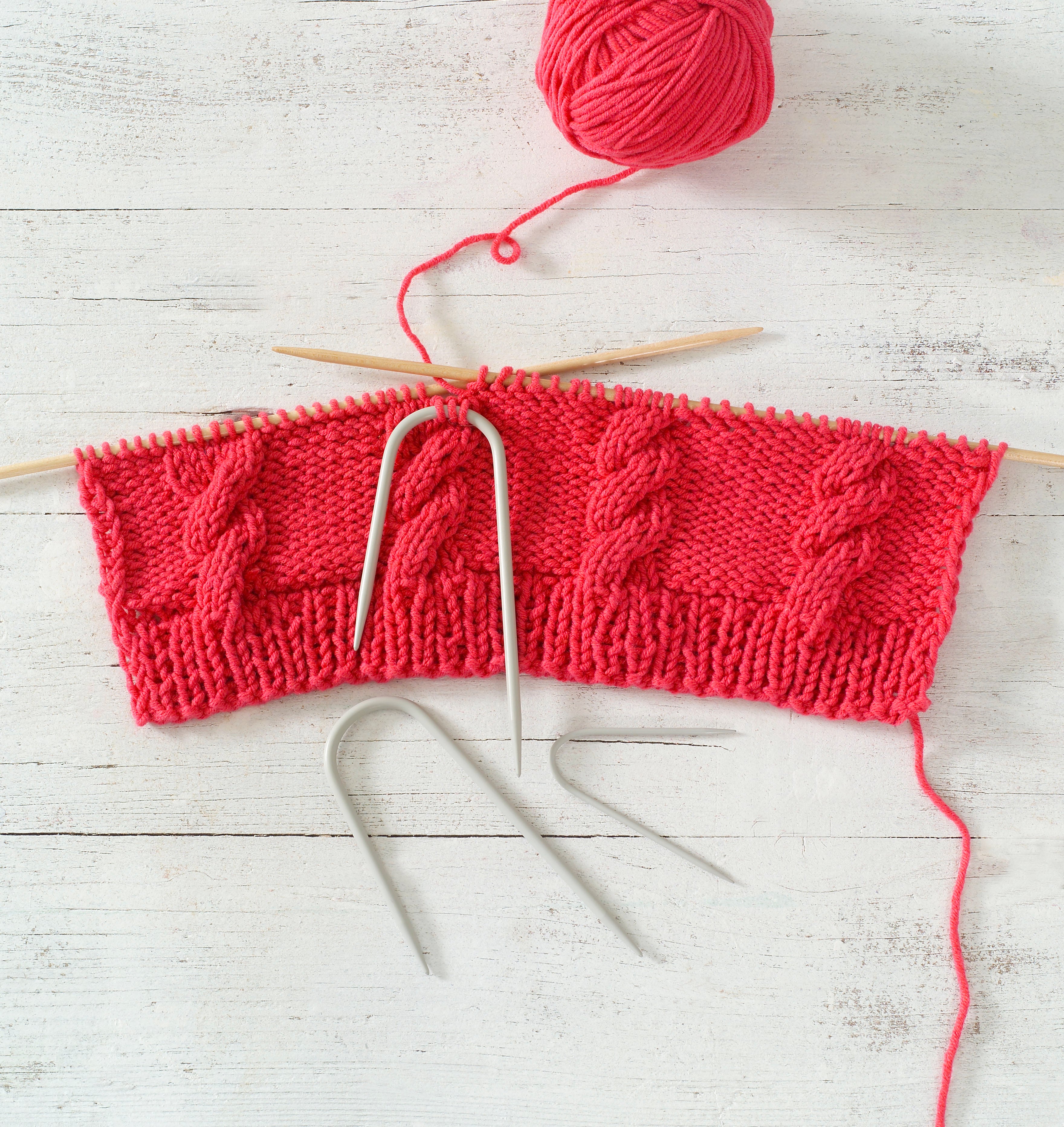 Red knitted fabric with cable knitting needles on a light wooden surface