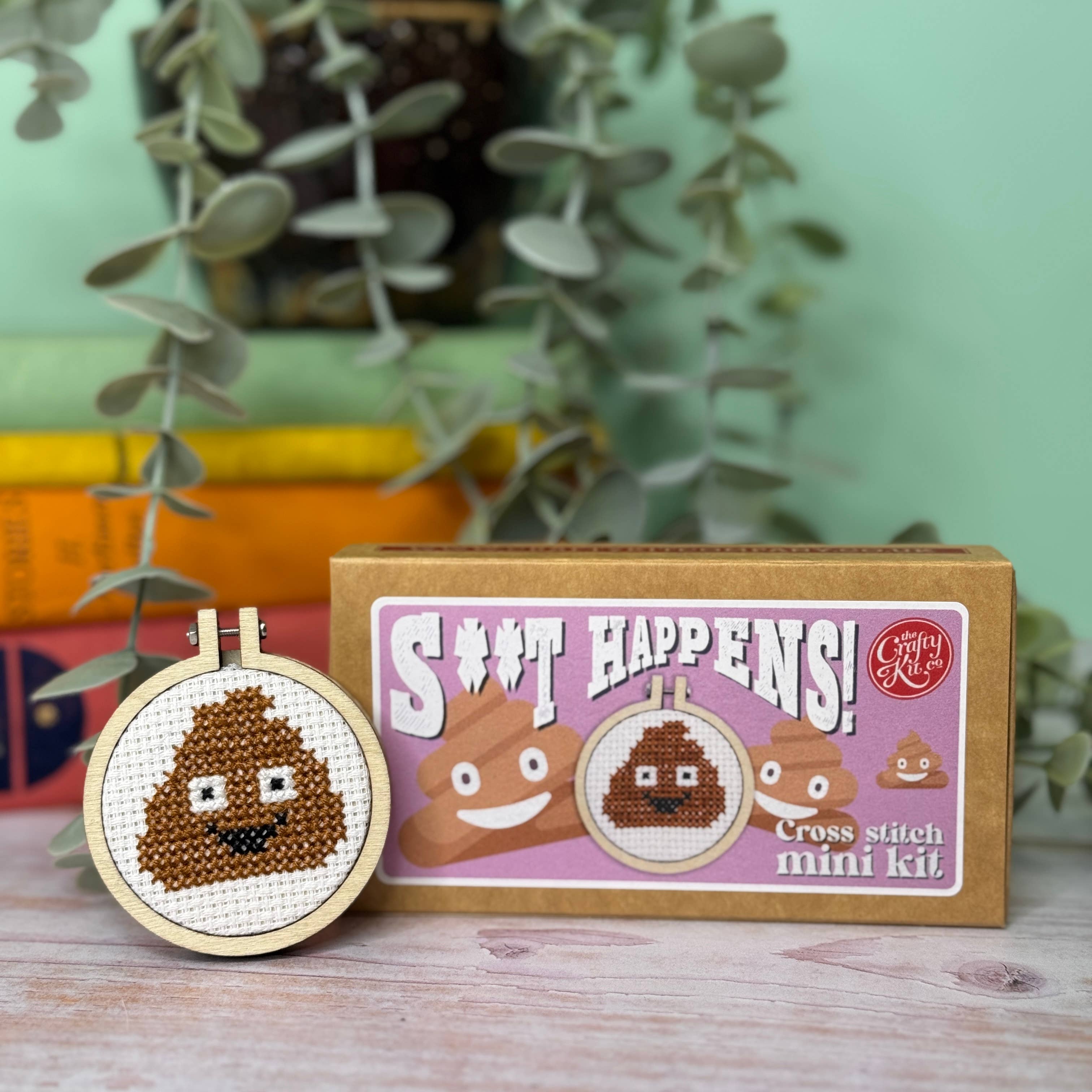 Cross stitch mini kit with poop emoji design and packaging labeled 'S#T HAPPENS!' on a wooden surface with plants in the background.