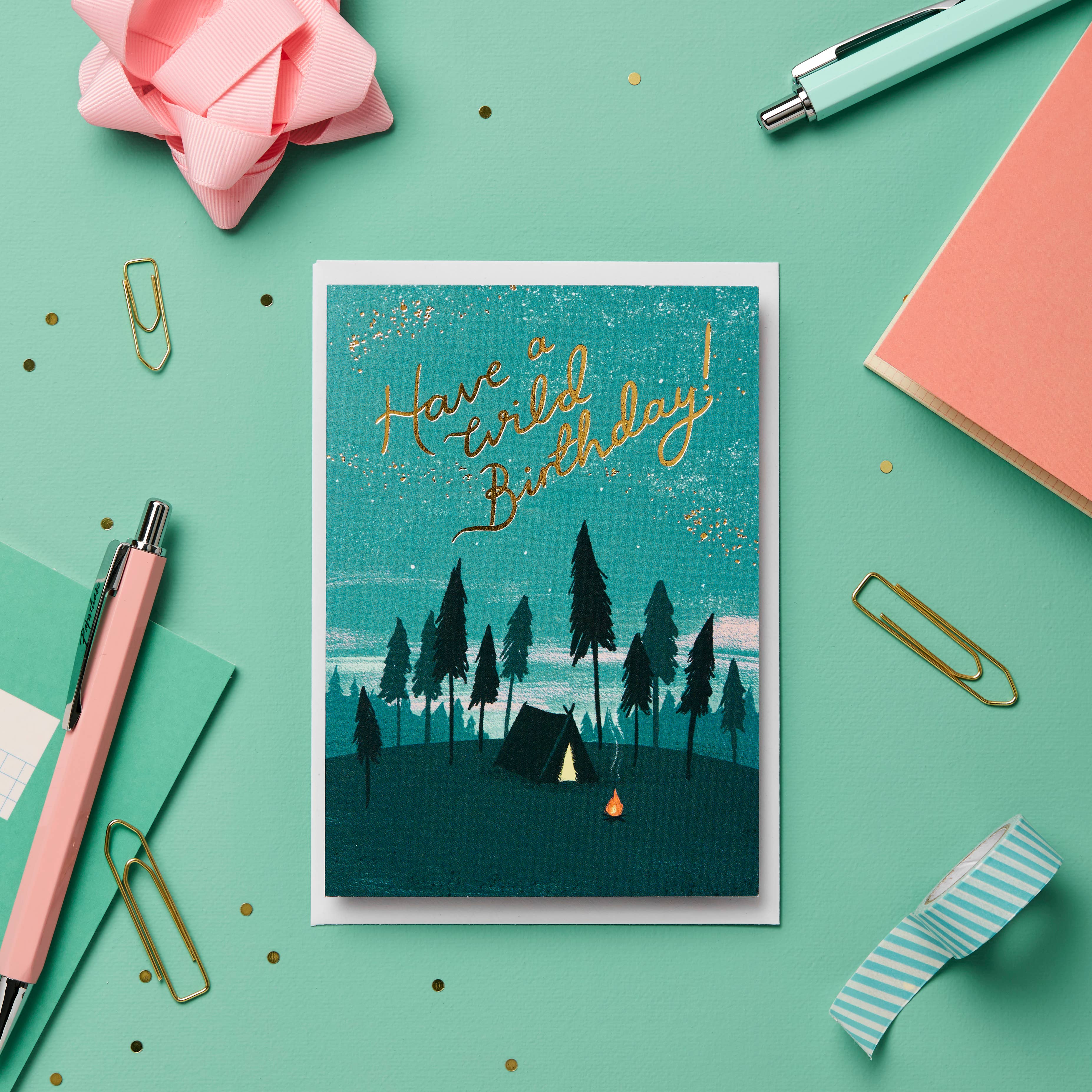 Greeting card with 'Have a wild birthday!' text on a teal background with camping design, surrounded by stationery items.
