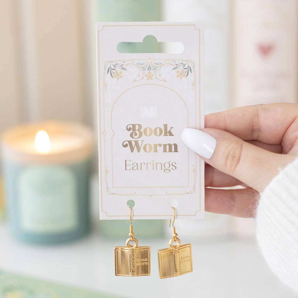Gold book-shaped earrings on a 'Book Worm' card with a blurred candle in the background