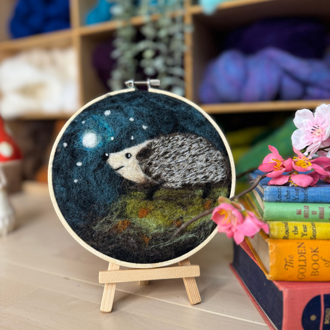 Embroidery hoop with a hedgehog design on a stand, surrounded by books and flowers.
