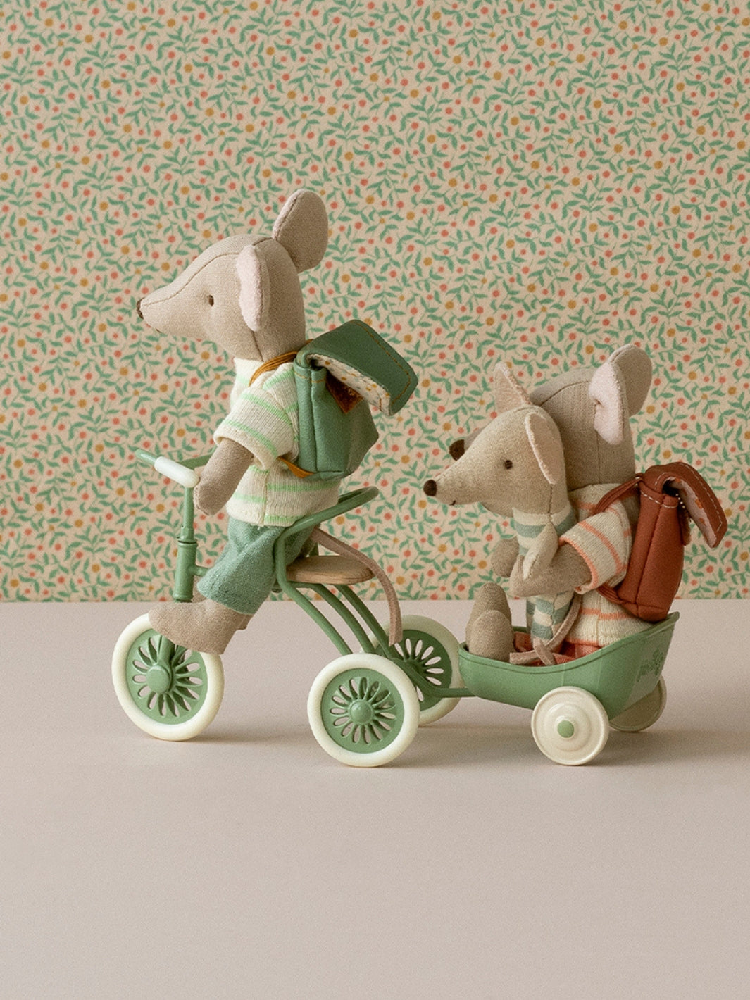 Two toy maileg mice on a tricycle and a cart against a patterned wall.