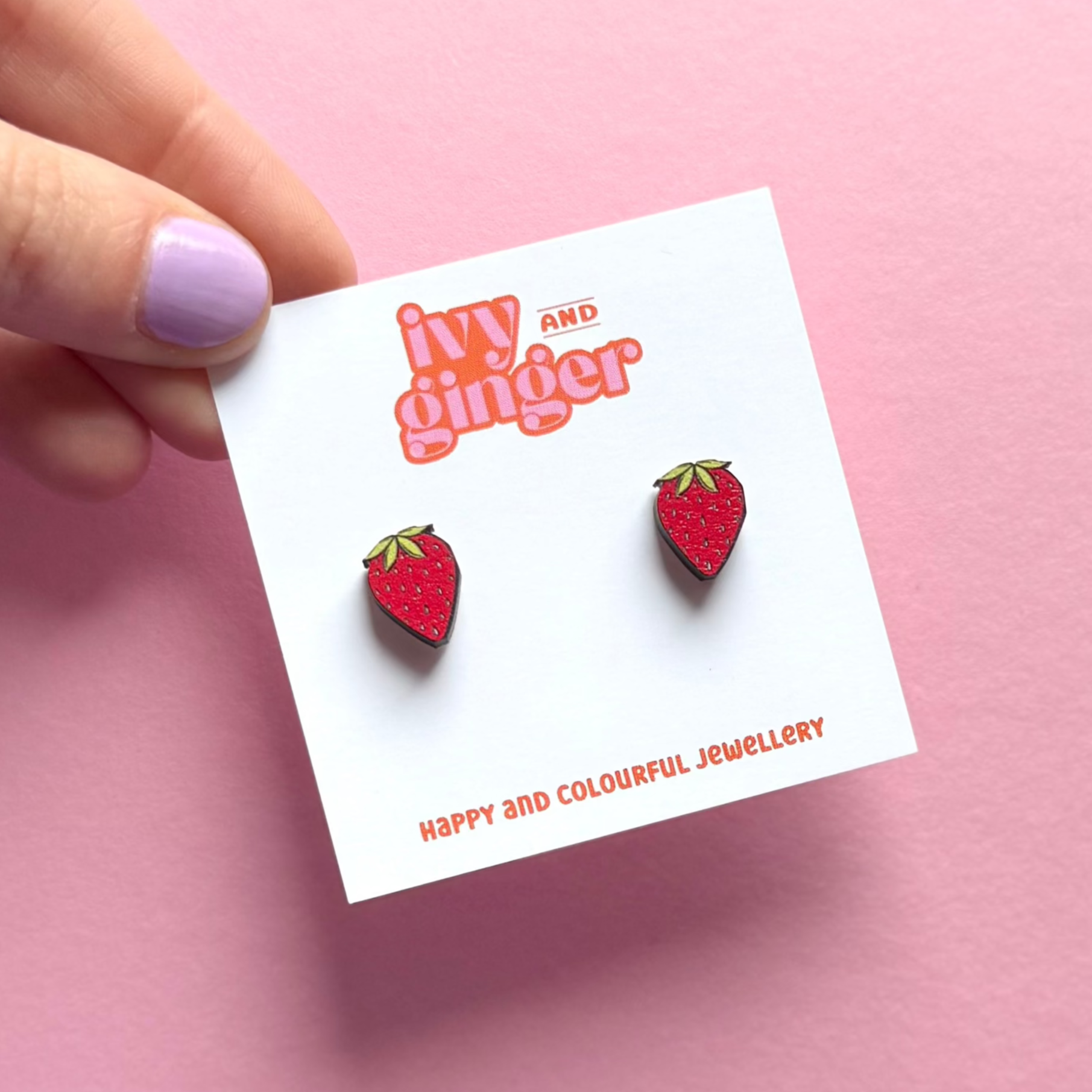 Strawberry-shaped earrings on a 'ivy and ginger' branded card against a pink background