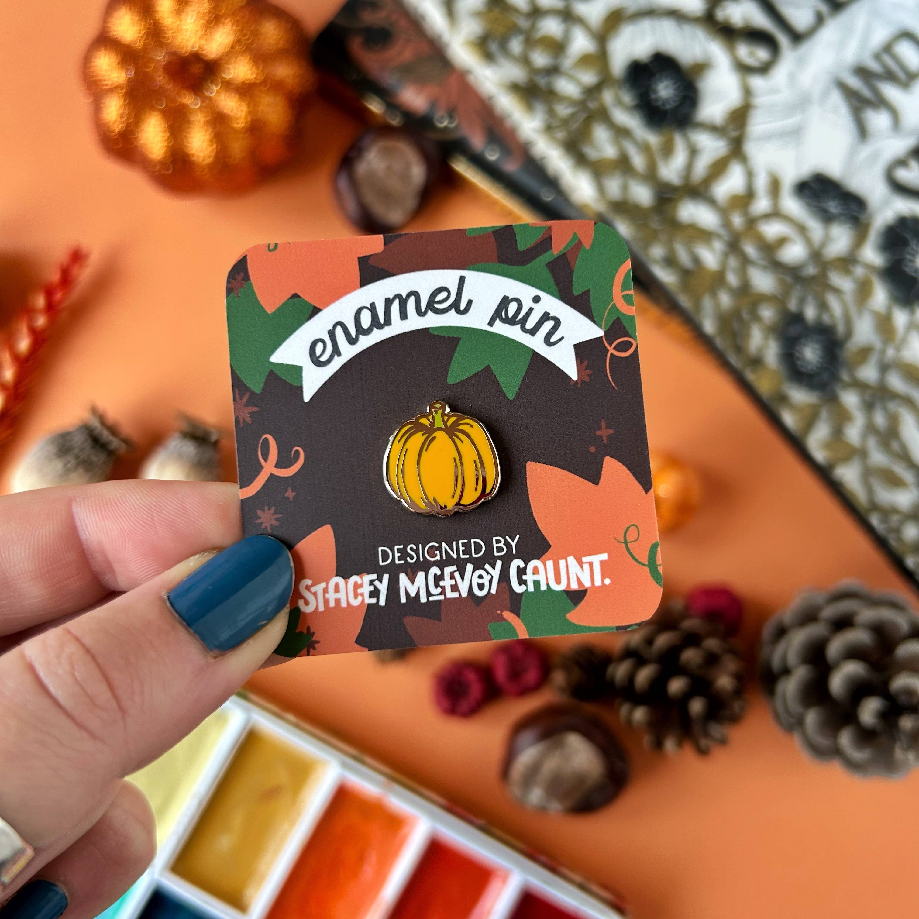 Enamel pin with pumpkin design on a card, held by a hand with blue nail polish, against a decorative background.