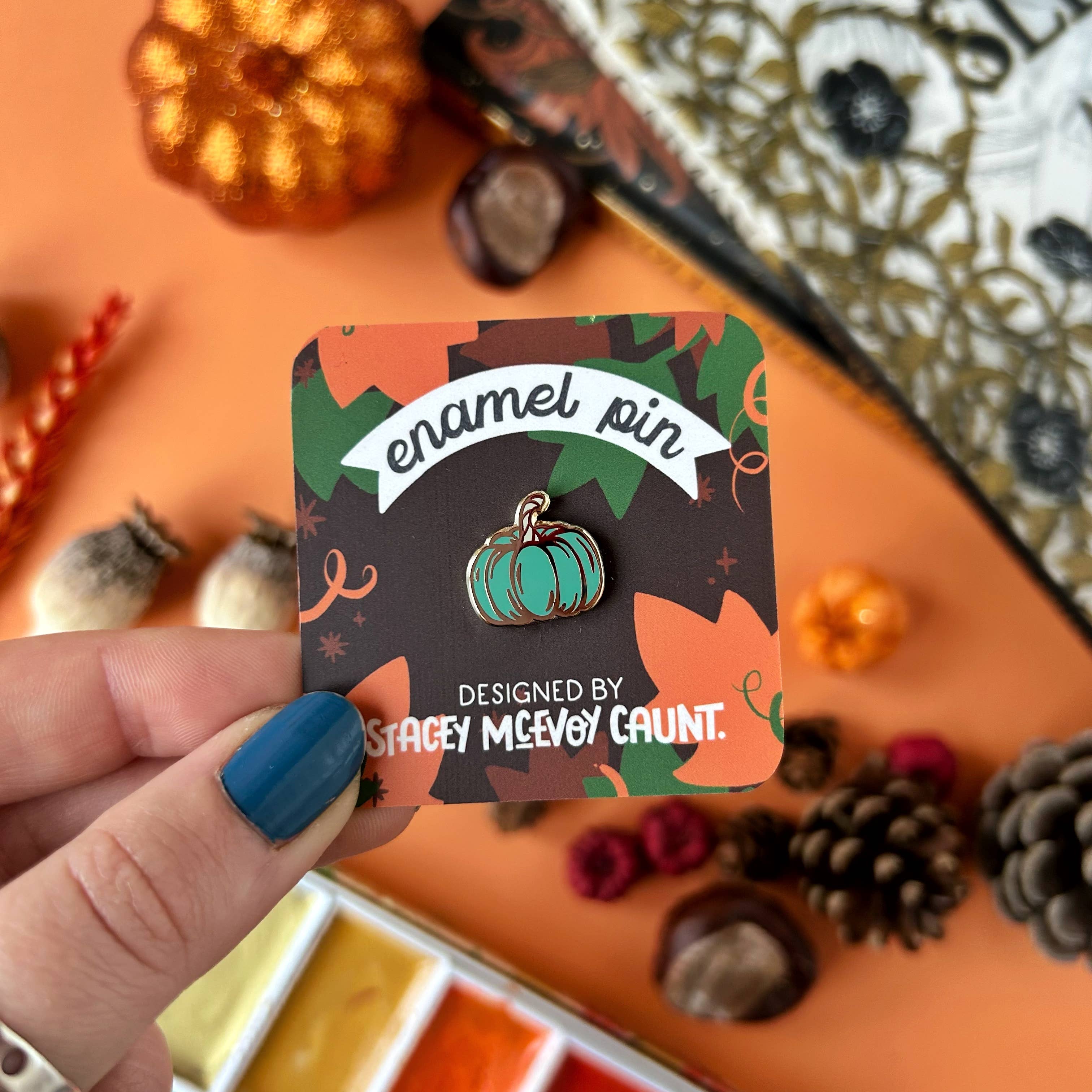 Enamel pin with decorative packaging held by a hand against a Halloween-themed background.
