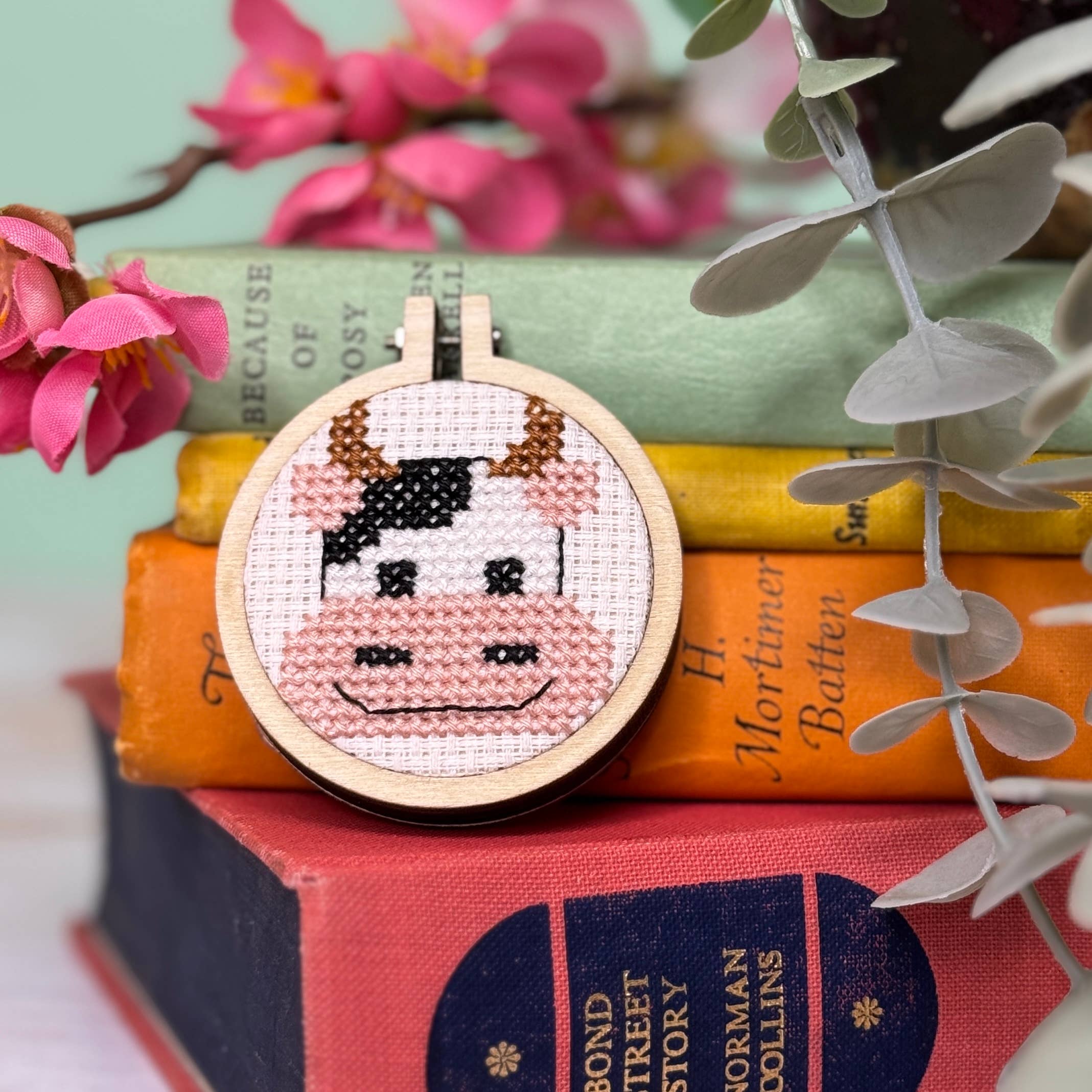 Cross-stitched cow design on a wooden hoop, placed on books with flowers and leaves in the background.