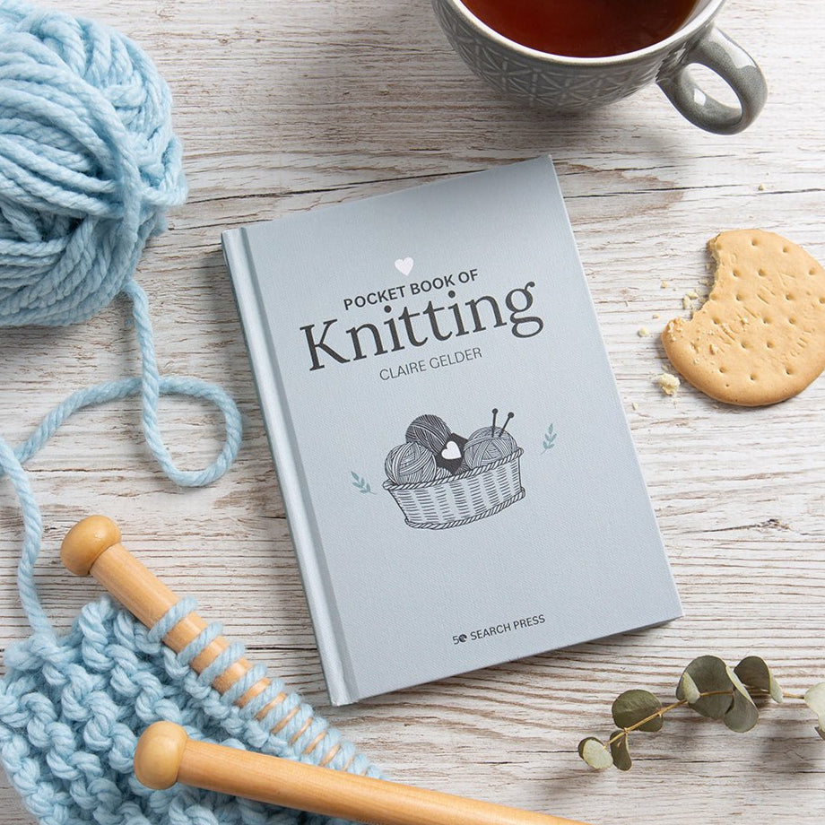 Knitting book titled 'Pocket Book of Knitting' by Claire Gelder on a wooden surface with yarn, knitting needles, and a cup of tea.