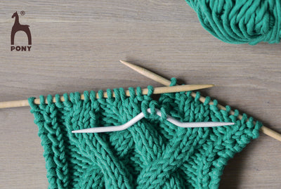 Green yarn being knitted with wooden needles on a wooden surface, featuring the Pony brand logo.