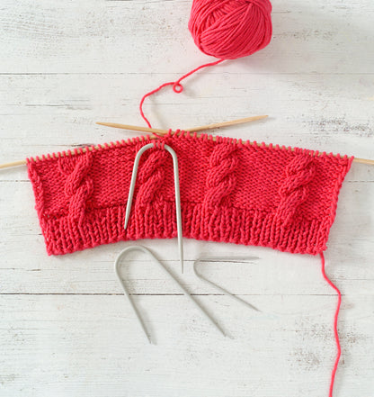 Red knitted fabric with cable knitting needles on a light wooden surface