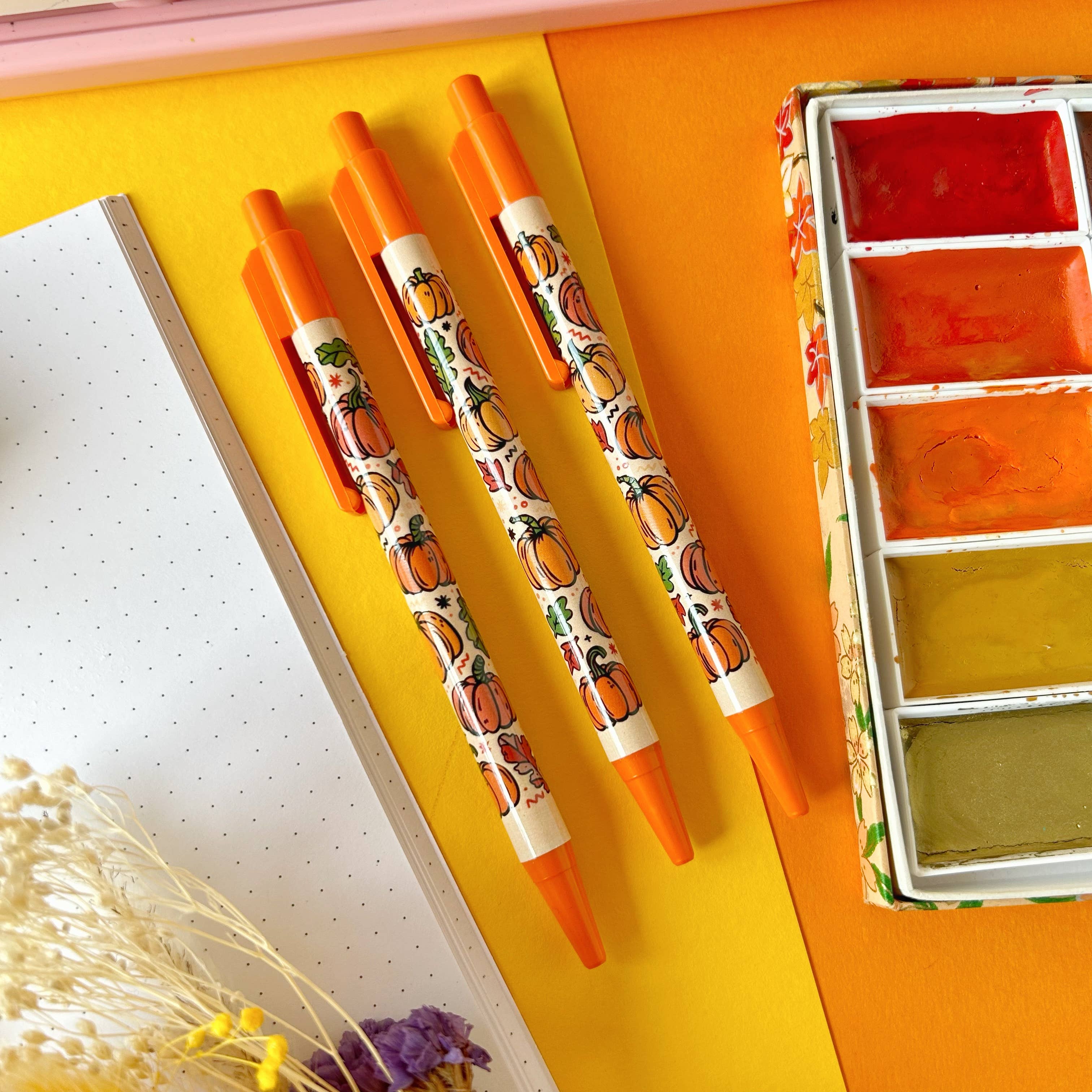 Pumpkin-themed pens on a yellow surface with a watercolor set.