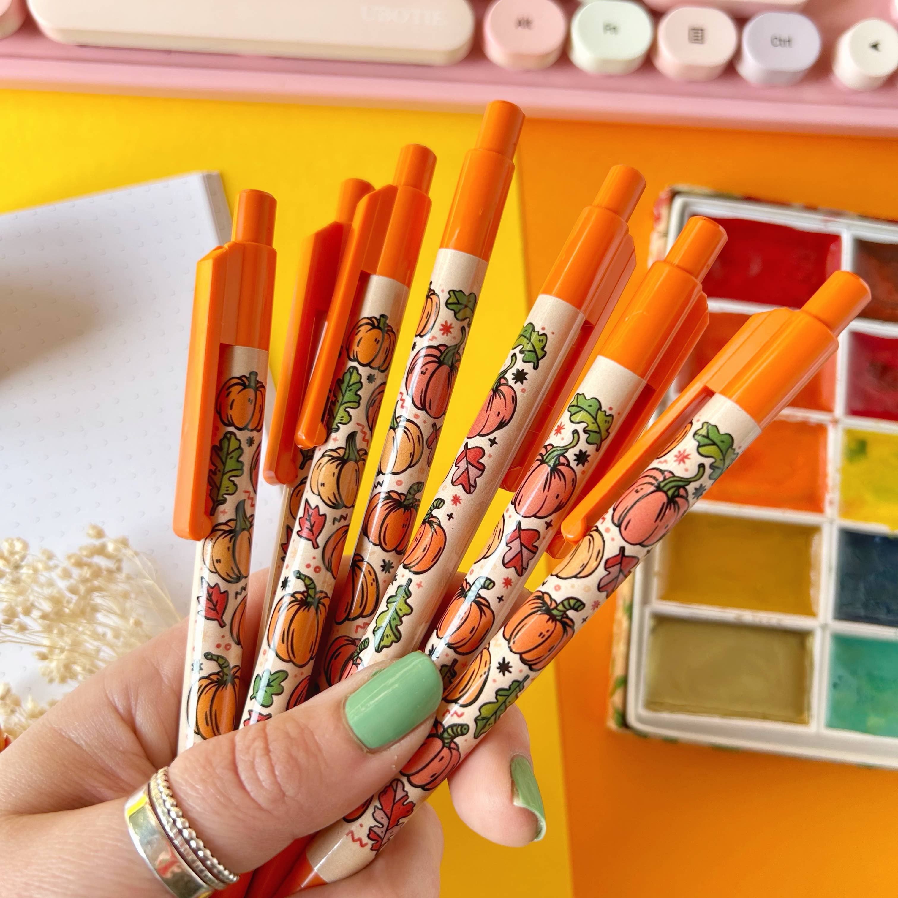 Hand holding pens with pumpkin designs on a colorful background
