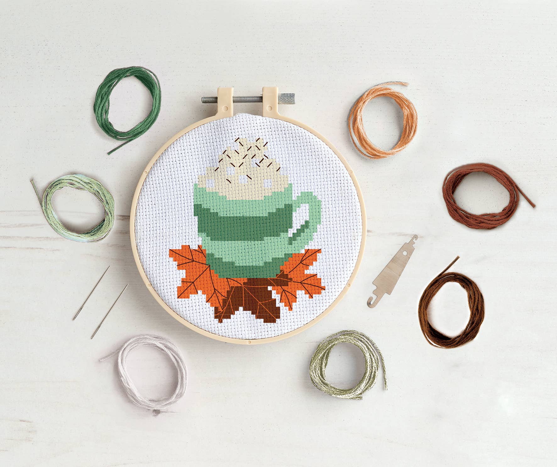 Cross-stitch hoop with a green mug design surrounded by embroidery threads on a light background