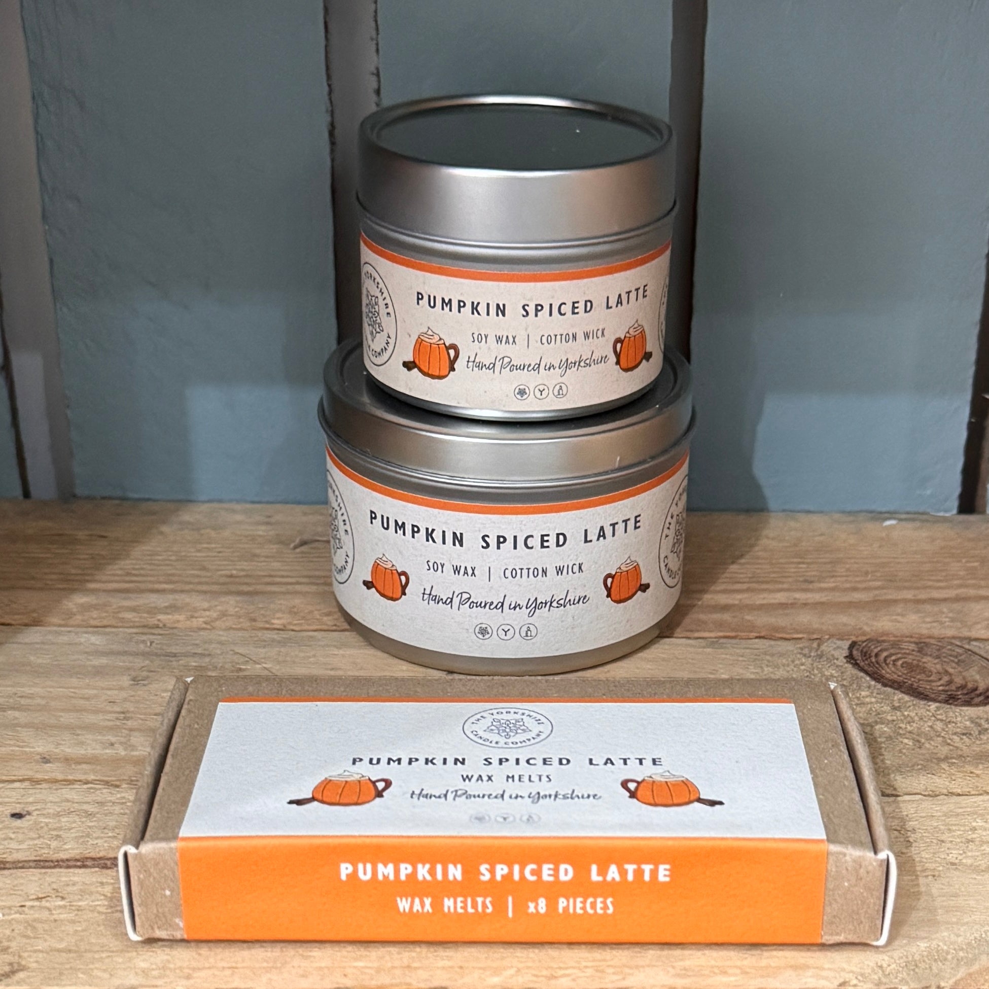 Two tins of 'Pumpkin Spiced Latte' candles stacked on a wooden surface with packaging in the foreground.