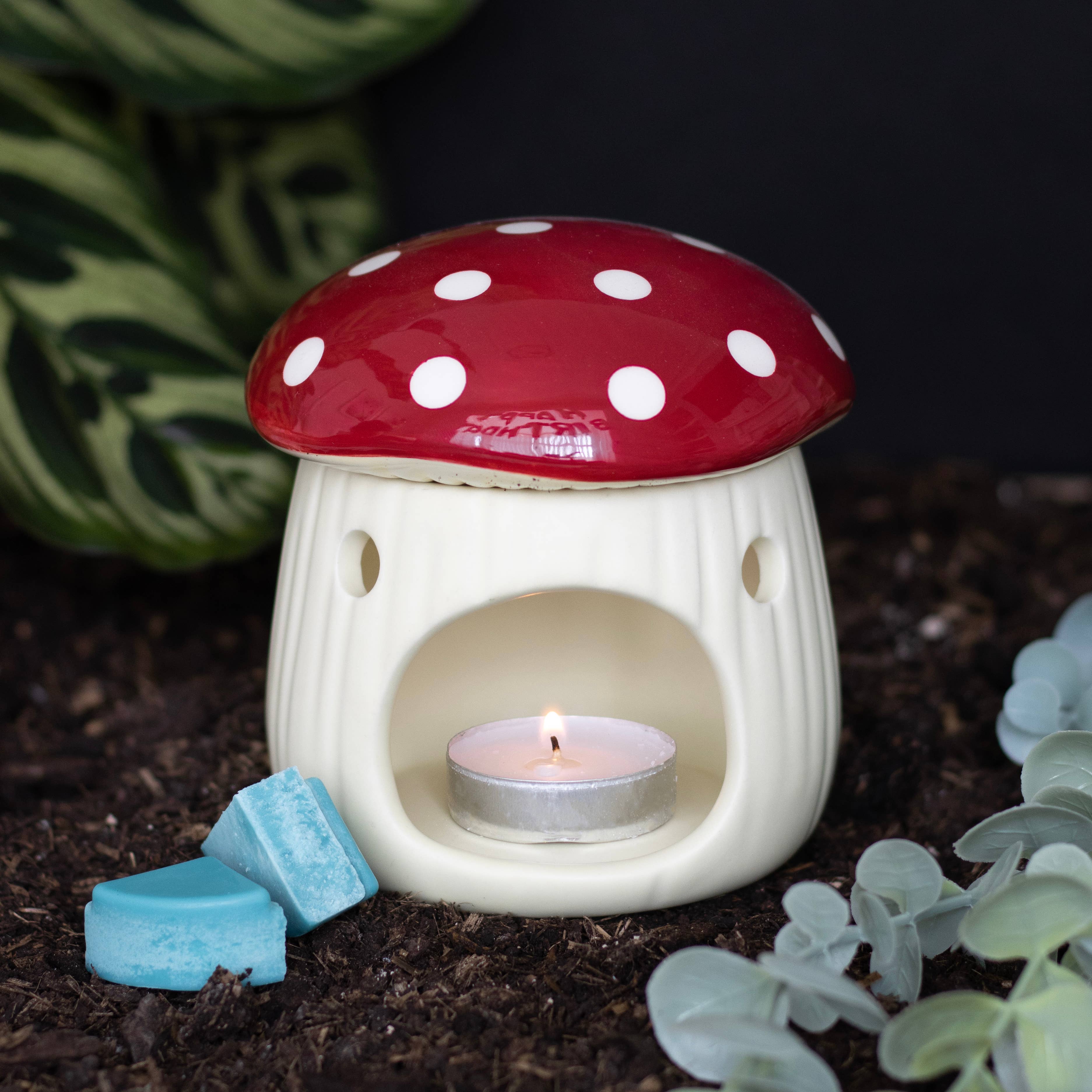 Mushroom-shaped ceramic oil burner with a lit candle inside, surrounded by plants on a dark background.