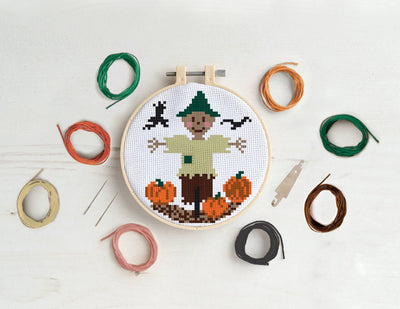 Cross-stitch hoop with a scarecrow design surrounded by colorful threads on a white background