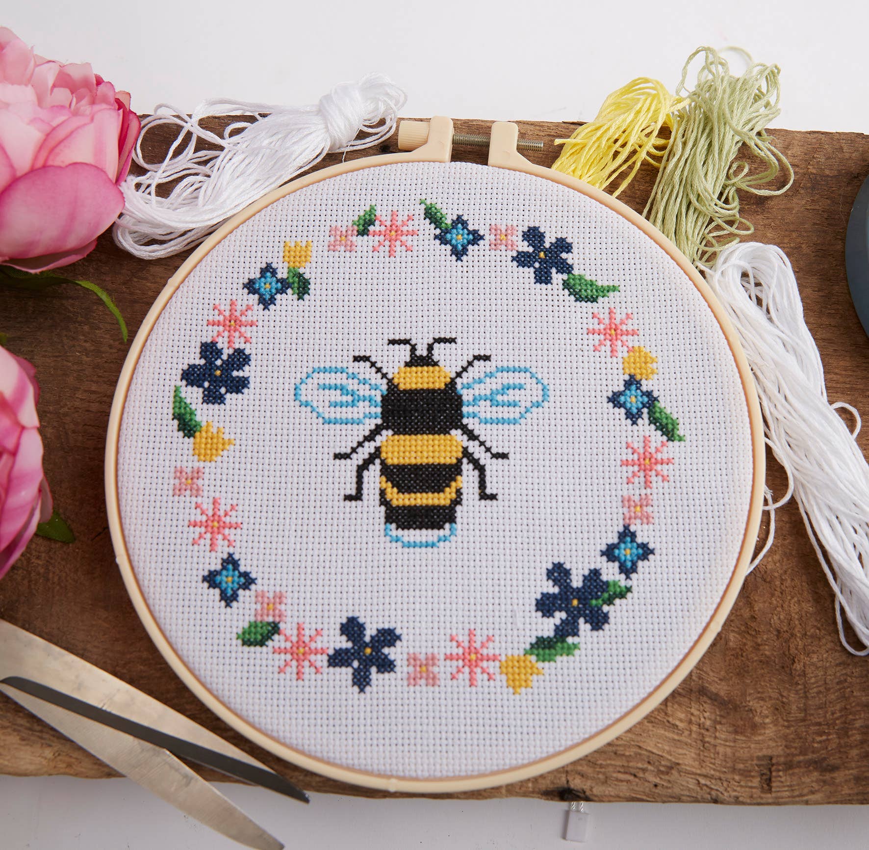 Embroidery hoop with a bee and floral design on a wooden surface with thread and scissors.