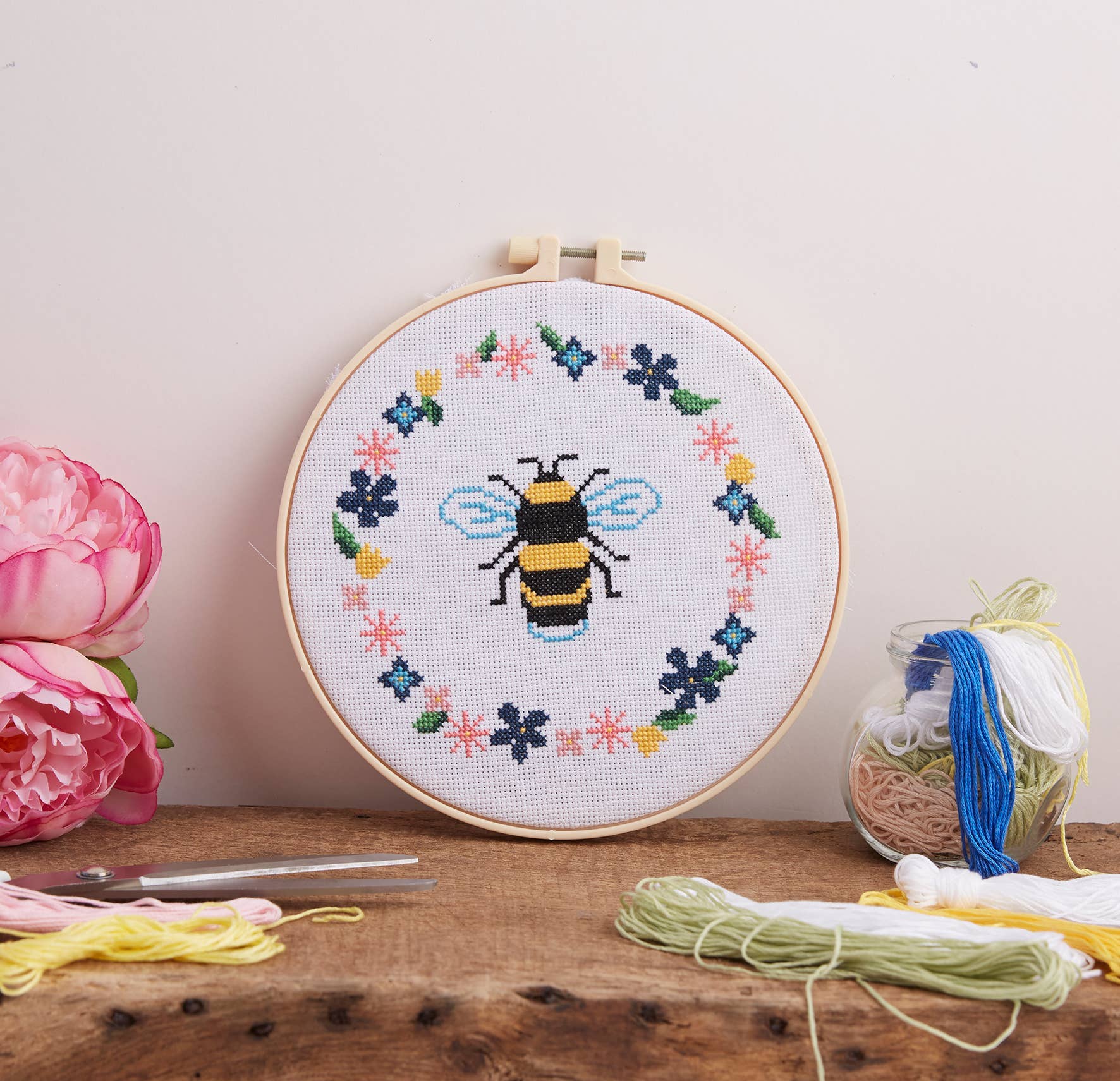 Embroidery hoop with a bee design surrounded by flowers on a wooden surface with embroidery supplies.