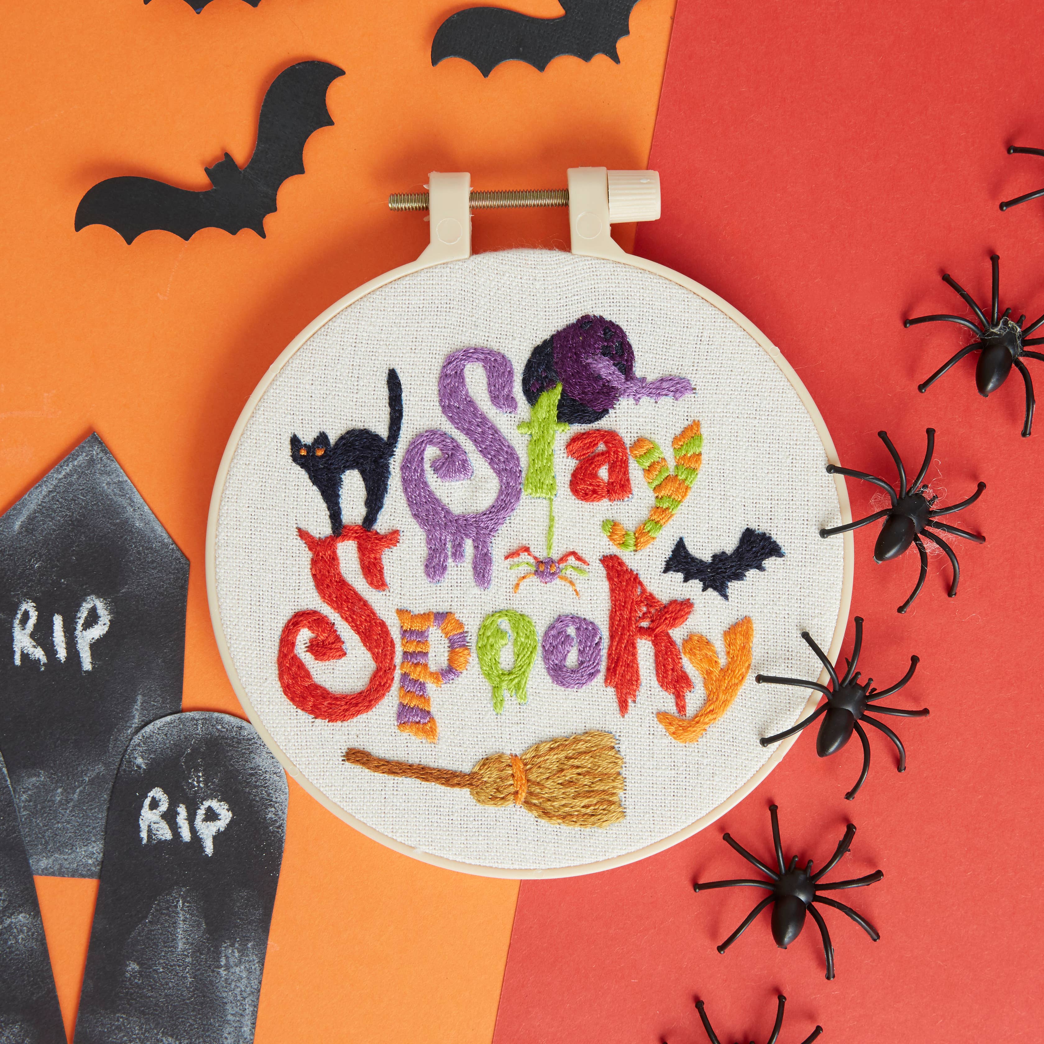 Embroidery hoop with 'Stay Spooky' text surrounded by Halloween decorations on a checkered background.