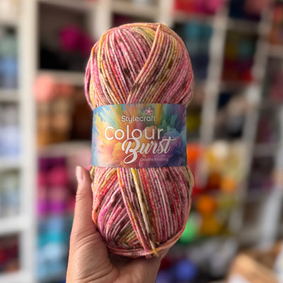 Multicolored skein of yarn with 'Stylecraft Colour Burst' label held in a hand, blurred shelves in the background.