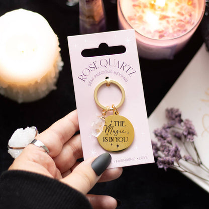 Keychain with 'The Magic is in You' message held by a hand, with packaging and candles in the background.