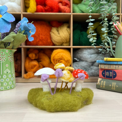 Felted mushroom sculptures on a green felt patch with colorful yarn and books in the background.